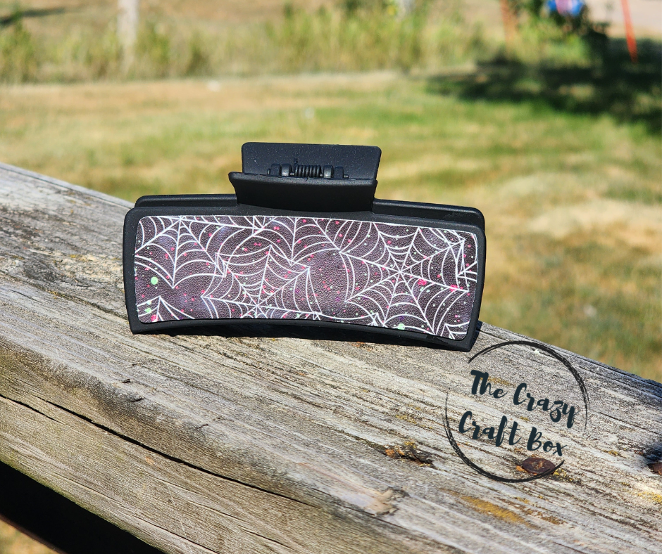 Whiskers n Bows black hair claw clip featuring a white spiderweb pattern and subtle glitter on a wooden fence.